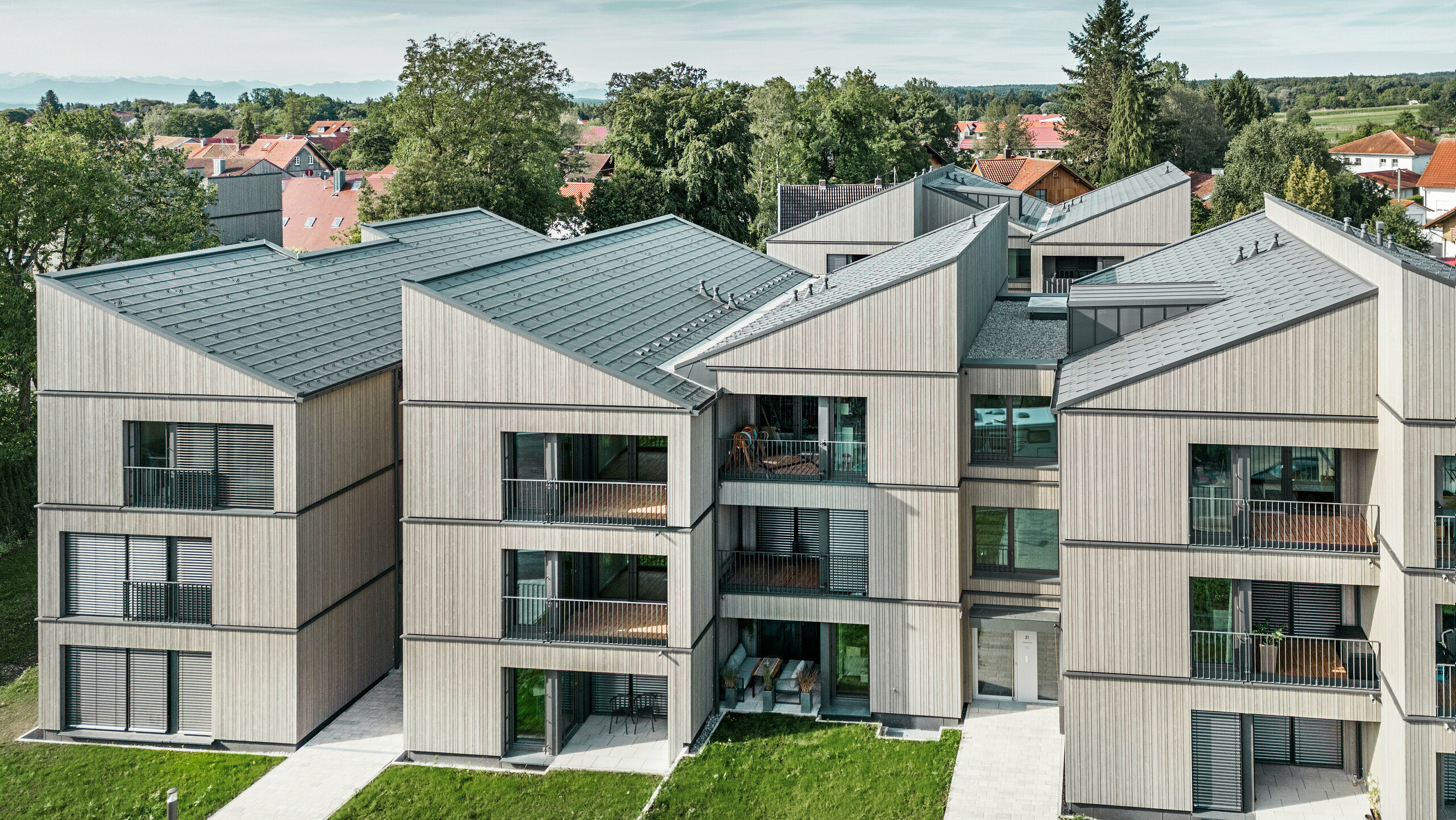 Luftaufnahme des modernen Mehrfamilienhauses am Schmucker-Areal in Utting am Ammersee, mit nachhaltiger Holzhybridbauweise und eleganten Holzfassaden. Die Gebäude sind mit langlebigen PREFA Dachplatten R.16 in P.10 Dunkelgrau ausgestattet. Diese innovative Architektur, geplant von WWA Architekten aus München, zeichnet sich durch klare Linien und harmonische Integration in die grüne Umgebung aus. Die großzügigen Balkone und großen Fenster sorgen für eine helle und freundliche Wohnatmosphäre, während die hochwertigen Materialien für Langlebigkeit und Wetterbeständigkeit sorgen.