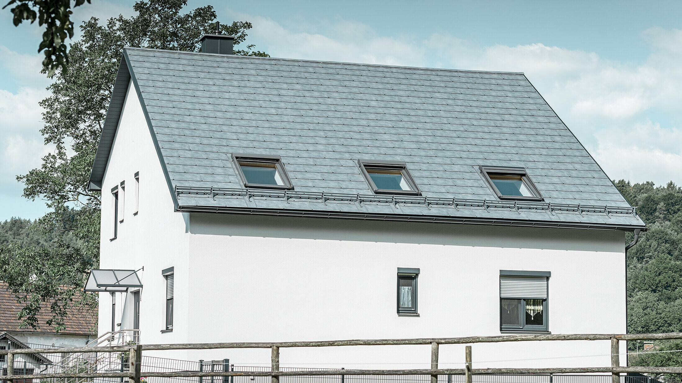 Das Satteldach des klassischen Einfamilienhauses wurde mit der neuen PREFA Dachplatte R.16 in Steingrau eingedeckt. In der Dachfläche wurden drei Dachfenster eingefügt und Schneerechen montiert. Die Fassade ist einfach in Weiß gehalten.