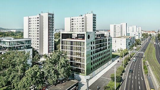 Pogled iz zraka na stanovanjsko in poslovno stavbo „Pick-Nick“ na trgu Straßburger Platz v Dresdnu s presenetljivo fasado PREFA iz aluminijastih rombov v različnih odtenkih zelene in bež barve. Stavba je vpeta v sodobno urbano okolje z visokimi stavbami in zelenimi površinami ter stoji ob glavni cesti. Presenetljiva arhitektura in visokokakovostna fasada PREFA predstavljata vizualne poudarke v mestni krajini.