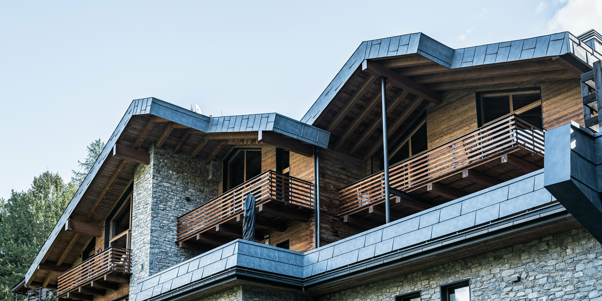 Detailný pohľad na moderný bytový dom v Courmayeur s balkónmi a kombináciou stien z prírodného kameňa a dreveného obkladu. Výrazné strešné a fasádne panely z hliníka PREFA vo farbe P.10 kamenná šedá zdôrazňujú vysoko kvalitnú architektúru. Konštrukcia je rámovaná okolitými borovicami, ktoré vytvárajú prirodzenú a pokojnú atmosféru a podčiarkujú harmonickú súhru moderných a tradičných stavebných prvkov.