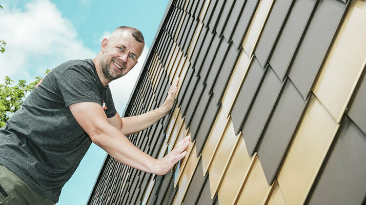 PREFA Handwerker mit grauem T-Shirt an einer PREFA Rauten-Fassade in Braun und Mayagold.