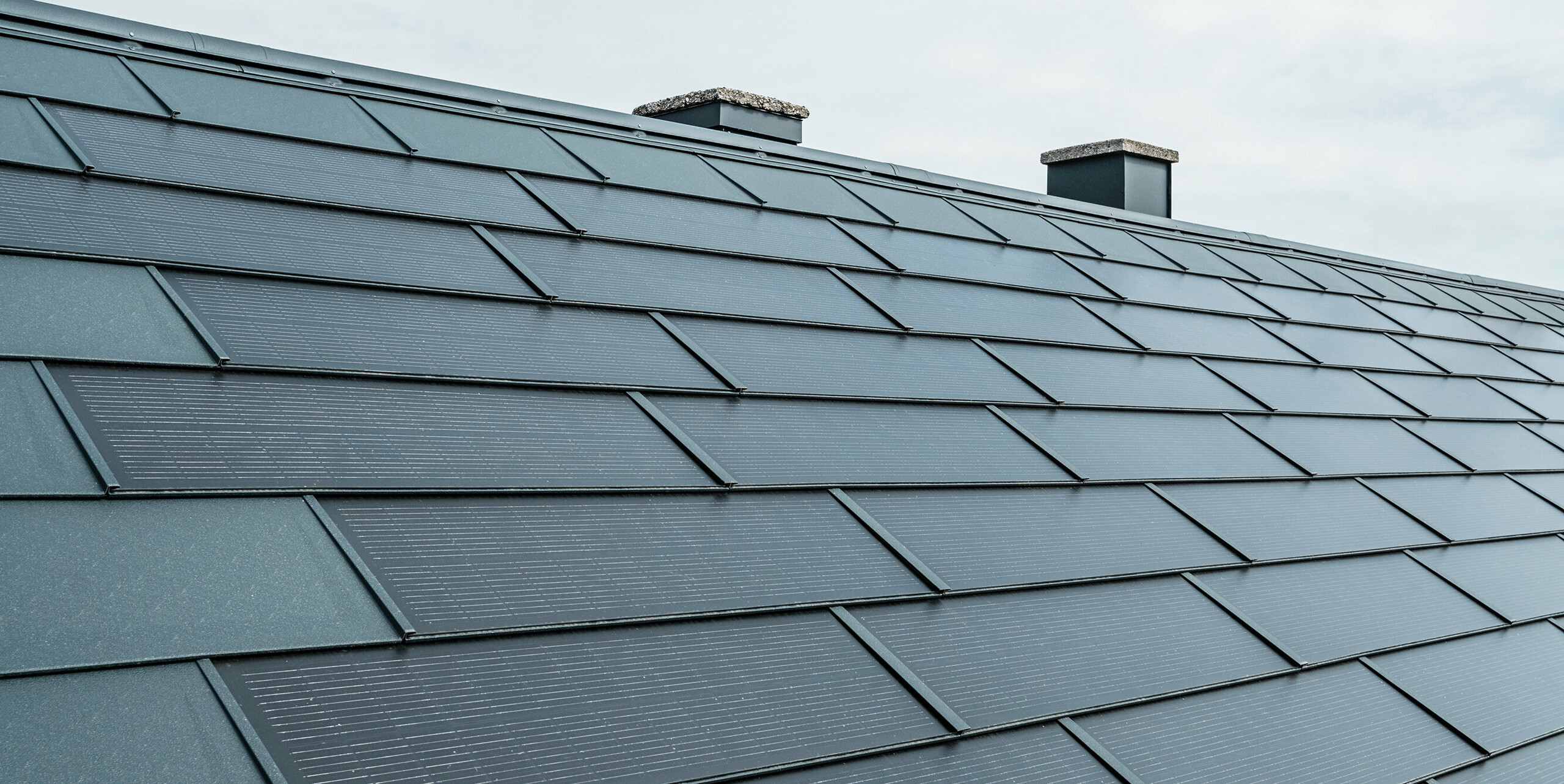 Perspektivni pogled na streho, opremljeno s PREFA solarnimi strešnimi ploščami v antracitni barvi. Plošče so natančno položene in zaradi vgrajenih fotovoltaičnih celic ponujajo inovativno rešitev za pridobivanje energije. V ozadju se dvigata dva dimnika, ki ju prekinja oblačno nebo, kar poudarja kombinacijo tradicionalne strešne arhitekture in sodobne sončne tehnologije.