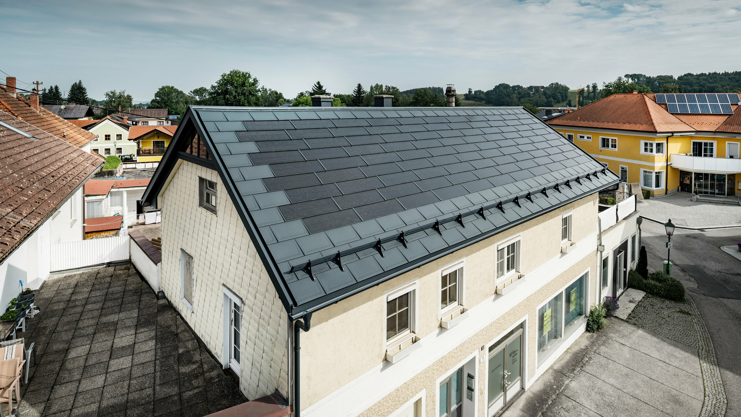 Pogled od zgoraj na enodružinsko hišo v Mettmachu v Avstriji. Starejši objekt je bil prenovljen s PREFA solarnimi strešnimi ploščami v antracitni barvi. Sistemi za zaščito pred snegom in jasne linije solarnih strešnih plošč poudarjajo čisto izdelavo aluminijastih izdelkov PREFA. V ozadju je v kontrastu s streho sosednje stavbe, ki je obarvana oranžno. Fotonapetostni moduli so nameščeni na strehi sosednje enodružinske hiše v klasični strešni rešitvi, kar nakazuje na pestrost energijsko učinkovite izvedbe streh.