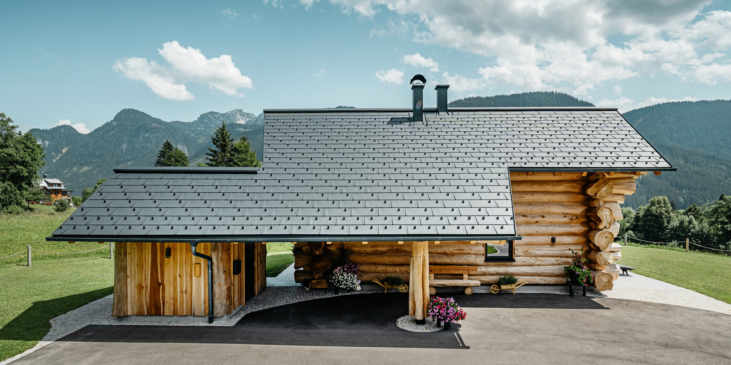 Stranski pogled na vabljivo leseno hišo v kraju Gosau v Avstriji, ki jo odlikuje robustna aluminijasta streha PREFA s strešnimi ploščami R.16 v P.10 antracitni barvi. Slika prikazuje harmonično povezavo med tradicionalno gradnjo iz hlodov in moderno, kakovostno strešno kritino, ki odraža dolgo življenjsko dobo in estetski dizajn aluminijastih izdelkov PREFA. Lesena hiša stoji na ozadju zelenih gričev in veličastne gorske panorame, ki poudarja naravno okolje.