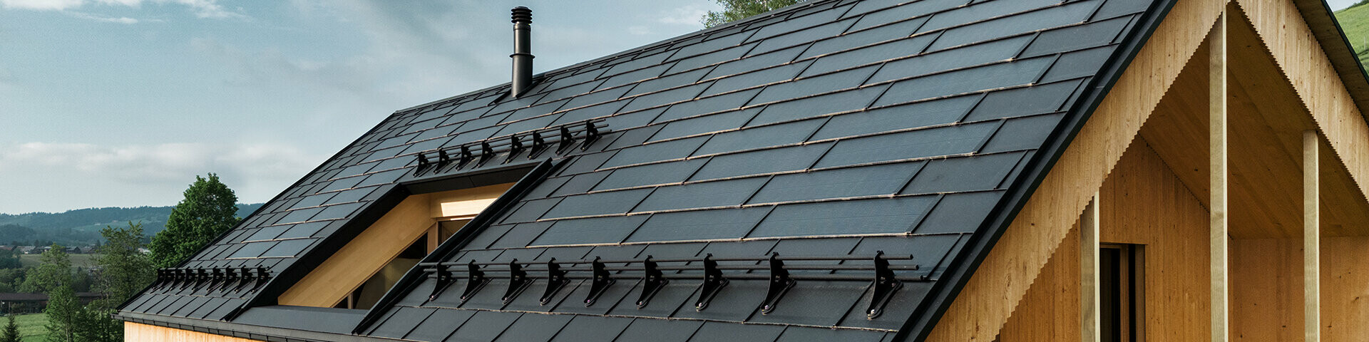 Solarna strešna plošča PREFA v črni barvi na eko hiši v Eggu v Avstriji - trpežna, energetsko učinkovita in estetska za sodobno arhitekturo. Inovativna solarna strešna plošča združuje streho, odporno proti nevihtam, s fotovoltaičnim sistemom. Vgrajeni fotovoltaični moduli se harmonično zlijejo s streho samostojne hiše.