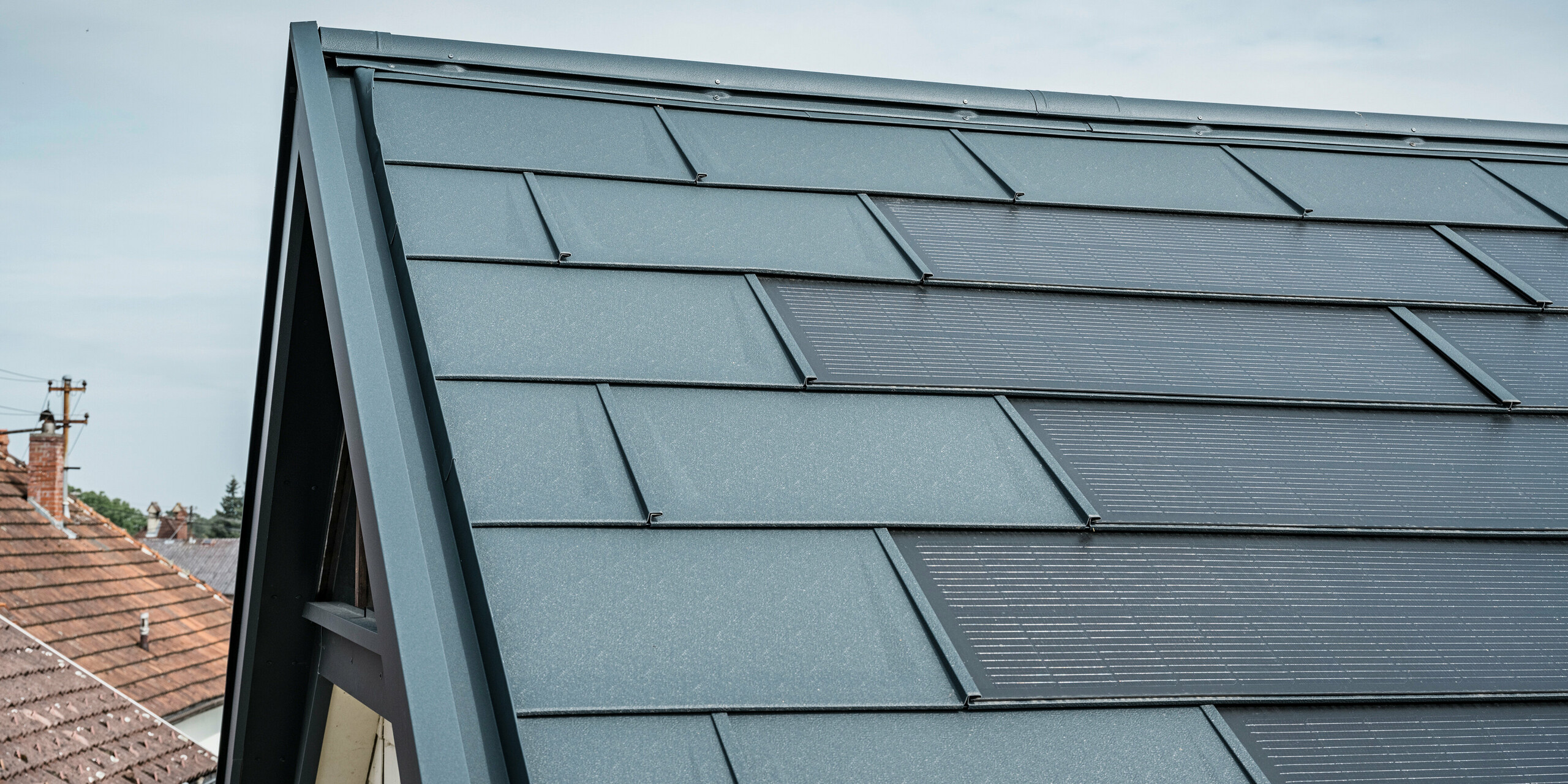 Podroben pogled na streho hiše, ki je opremljena z inovativno PREFA solarno strešno ploščo. Strešne plošče z integriranimi fotovoltaičnimi celicami so v elegantni antracitni barvi. Homogena površina se brezhibno zliva s streho in tako ustvarja moderen in čist videz. Inovativni strešni sistem zagotavlja učinkovito rabo energije, ne da bi ob tem ogrožal estetiko.