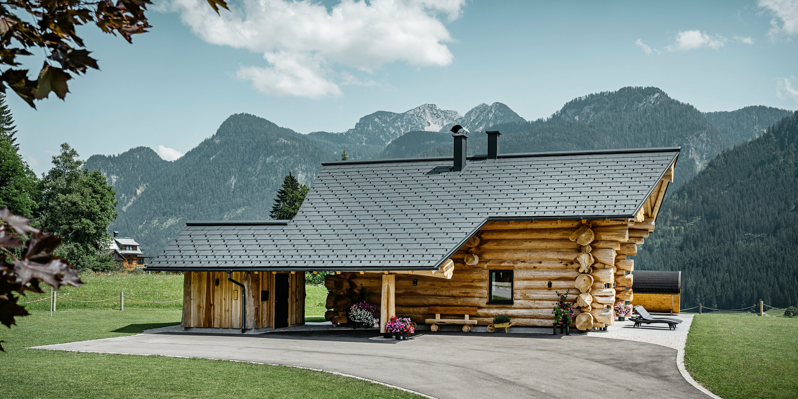 Brunarica v Gosau-u v Avstriji s strešnim sistemom PREFA v antracitni barvi. Preizkušene strešne plošče R.16 ustvarjajo privlačen kontrast naravnemu lesu brunarice. V ozadju je veličastna gorska panorama, ki poudarja mirno in slikovito lokacijo hiše. Podrobna fotografija prikazuje simbiozo tradicionalne avstrijske arhitekture in sodobne strešne tehnologije ter prikazuje robustno in estetsko kakovost aluminijastih izdelkov PREFA.