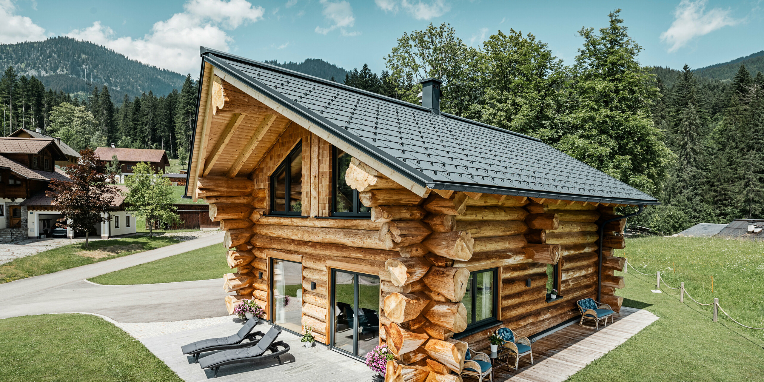 Rustikalna brunarica v kraju Gosau v Avstriji, opremljena s trpežnimi PREFA strešnimi ploščami R.16 v P.10 antracitni barvi. Aluminijaste strešne plošče majhnega formata so harmonično vpete v naravno pokrajino z bujno zelenimi drevesi in slikovitimi gorami v ozadju. Visokokakovostni strešni sistem v antracitni barvi je v močnem kontrastu s tradicionalnimi stenami iz lesenih hlodov in je optimiziran glede odpornosti na vremenske vplive in vzdržljivost. Ta slika ponazarja popolno kombinacijo tradicionalne gradnje in sodobnega dizajna PREFA.