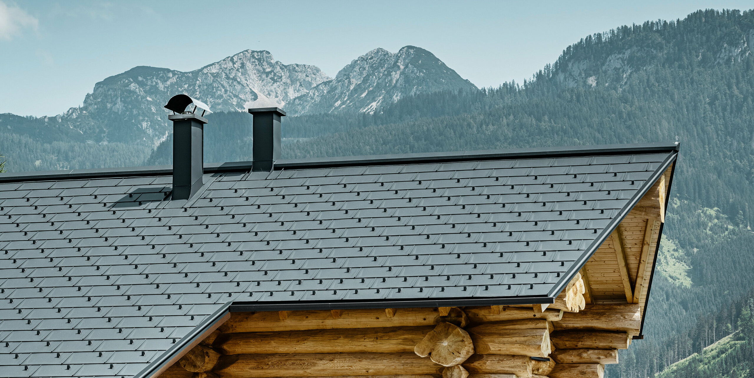Navdušujoč pogled na tradicionalno brunarico v Gosau-u v Avstriji pred impresivno gorsko panoramo. Hiša ima aluminijasto PREFA streho, sestavljeno iz strešnih plošč R.16 v P.10 antracitni barvi. Streha v antracitni barvi daje sodoben poudarek rustikalni leseni konstrukciji. Visokokakovostne strešne plošče se brezhibno prilegajo alpski pokrajini, medtem ko naravnost lesene hiše poudarja podeželski življenjski slog. Kombinacija naravnih materialov in estetske zasnove strehe daje tej nepremičnini brezčasen čar.