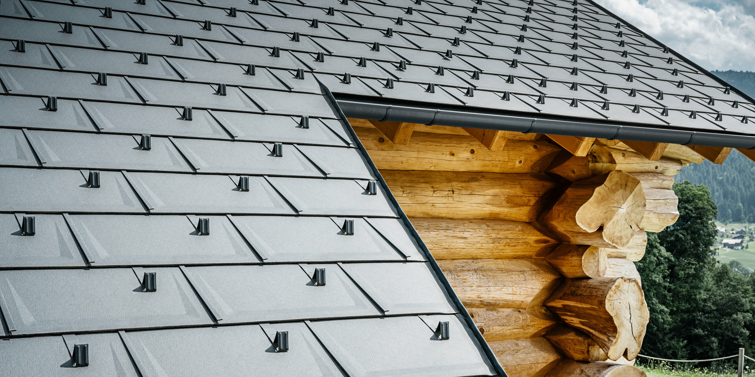 Bližnji pogled na streho s PREFA strešnimi ploščami R.16 v P.10 antracitni barvi na leseni hiši v Gosau-u v Zgornji Avstriji. Natančna obdelava in montaža robustnih strešnih plošč dokazujeta visoko kakovost aluminijastih izdelkov PREFA kot tudi popolno integracijo sodobnih aluminijastih gradbenih elementov v tradicionalne avstrijske tipe zgradb. V ospredju so vidni značilni snegobrani, ki zagotavljajo varnost v alpski zimi. Ta slika prikazuje harmonično prepletanje inovativnih strešnih sistemov PREFA z naravno estetiko lesene brunarice.