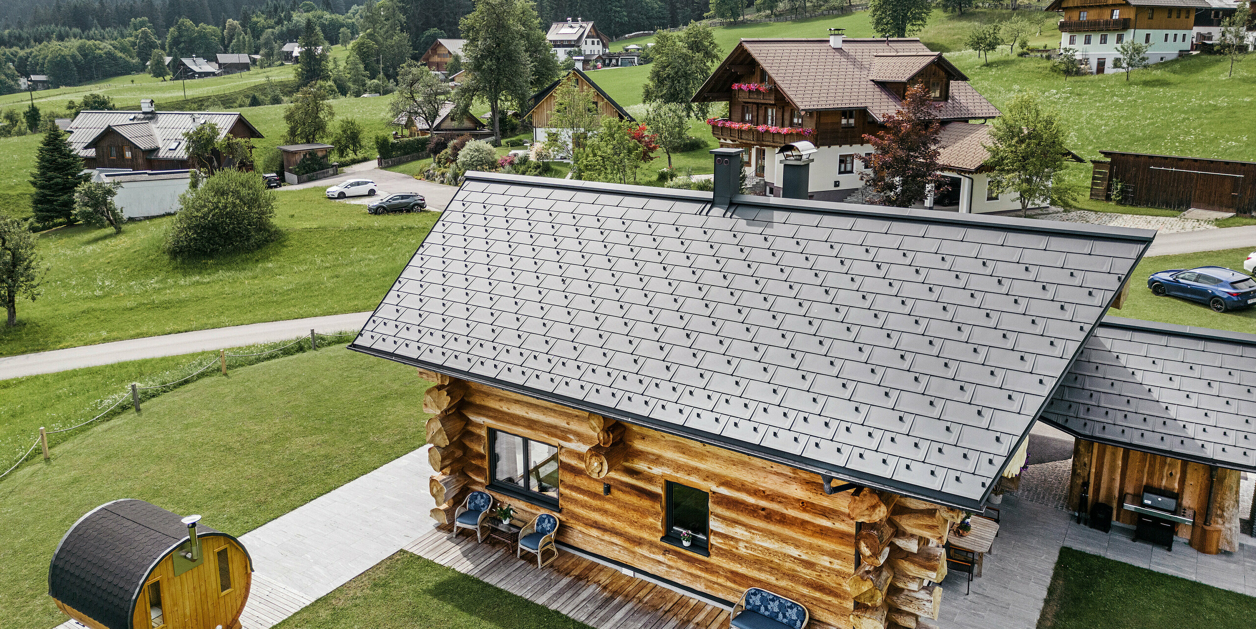 Pogled iz ptičje perspektive na rustikalno brunarico v kraju Gosau v Avstriji z visokokakovostno aluminijasto PREFA streho iz strešnih plošč R.16 v P.10 antracitni barvi. Streha kaže značilno strukturo in natančnost PREFA strešnih sistemov, ki se odlično ujemajo s podeželskim okoljem. Bujno zelenje travnikov, okoliške tradicionalne hiše in gozdnati griči v ozadju tvorijo naravni kontrast modernim strešnim elementom v antracitni barvi, ki v alpsko arhitekturo vnašajo trajnost in sodobno estetiko.