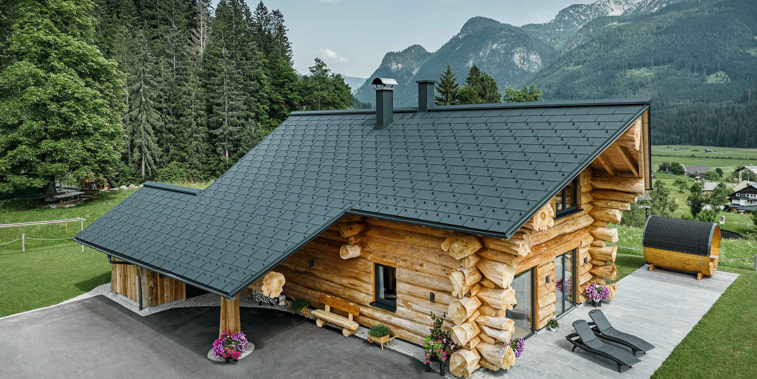 Poševni pogled na slikovito brunarico v Gosau-u v Avstriji, opremljeno s PREFA streho iz antracitnih strešnih plošč R.16. Arhitektura združuje rustikalni pridih brunarice s sodobnimi elementi, obdana z gostimi zelenimi gozdovi in impresivno gorsko kuliso. Hiša s svojo podeželsko eleganco in natančno položenimi aluminijastimi strešnimi elementi, ki so znani po svoji trpežnosti in odpornosti na vremenske vplive, izžareva vabljivo vzdušje. 