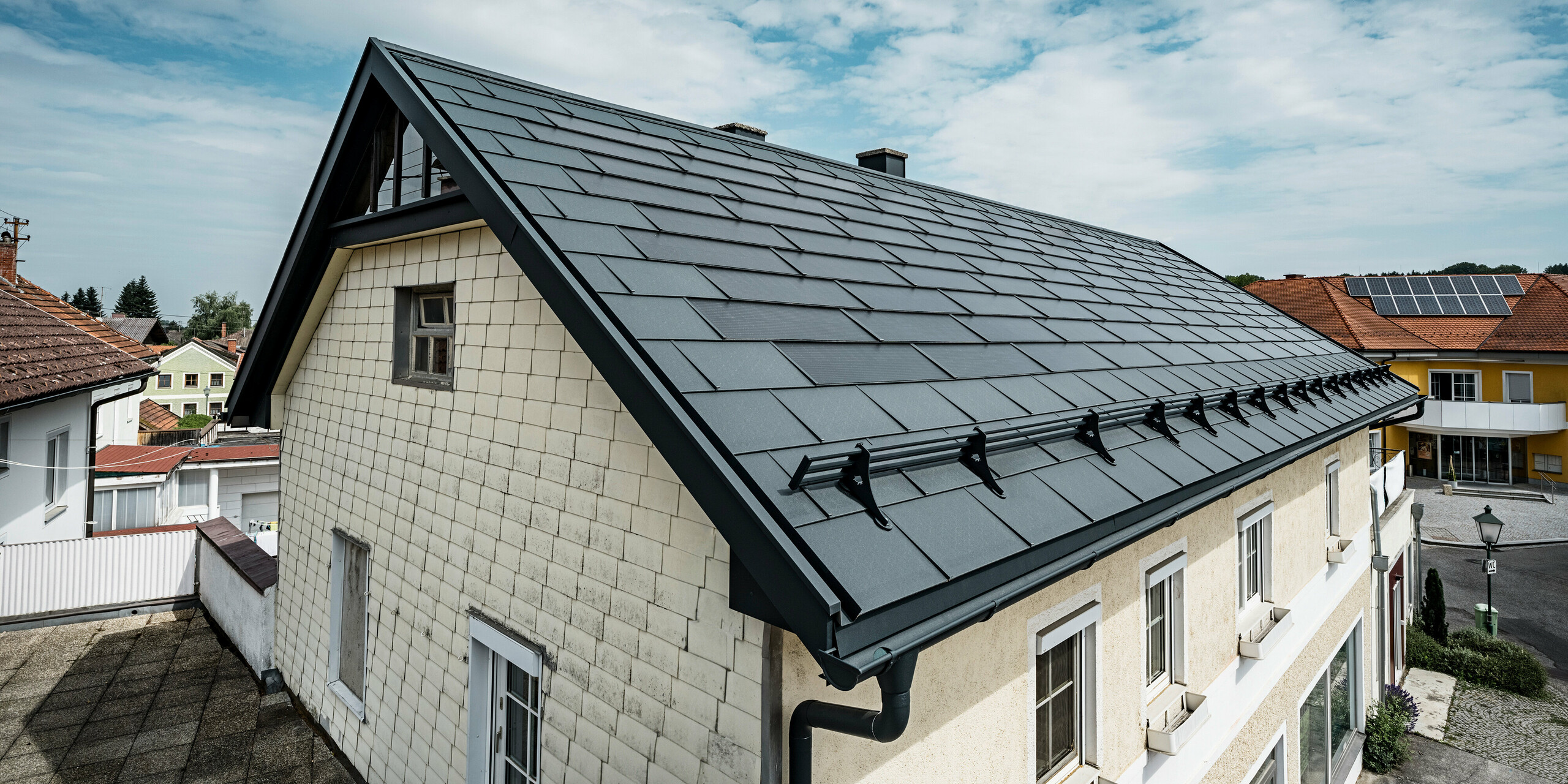 Pogled iz ptičje perspektive na streho enodružinske hiše v Mettmachu v Avstriji. Hiša je bila v okviru prenove opremljena s PREFA solarnimi strešnimi ploščami v antracitni barvi. Strukturirana površina strešnih plošč se ujema s preprosto eleganco hiše. Na rob strehe je pritrjen sistem snegobrana. V ozadju so vidne druge hiše s tradicionalnimi in solarnimi strešnimi rešitvami, kar poudarja sodobno, energetsko učinkovito zasnovo strehe v podeželskem okolju.