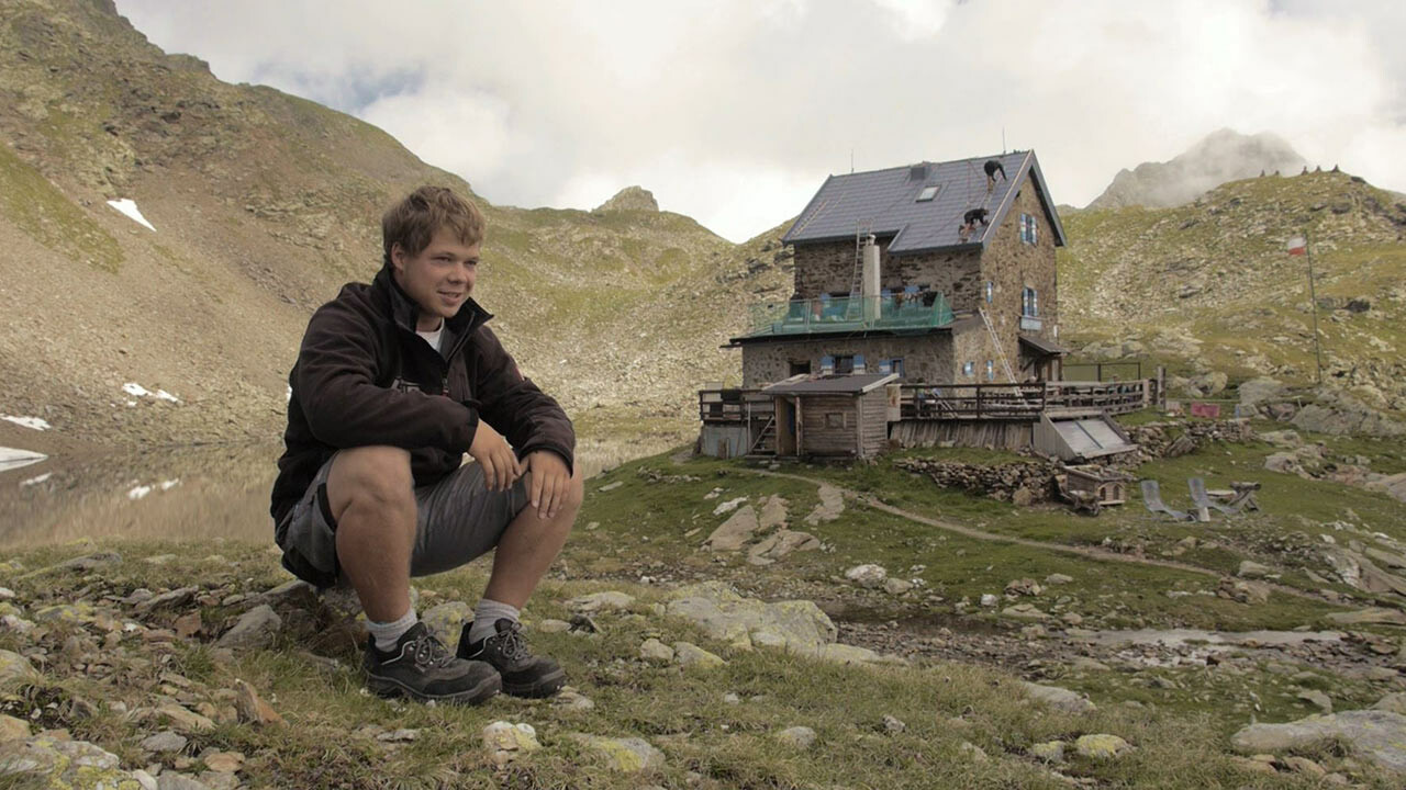 Pripravnik Jakob Schöttl sedi na skali. V ozadju je vidna gorska koča „Flagger Schartenhütte“, ki je bila na novo pokrita s strešno ploščo PREFA.