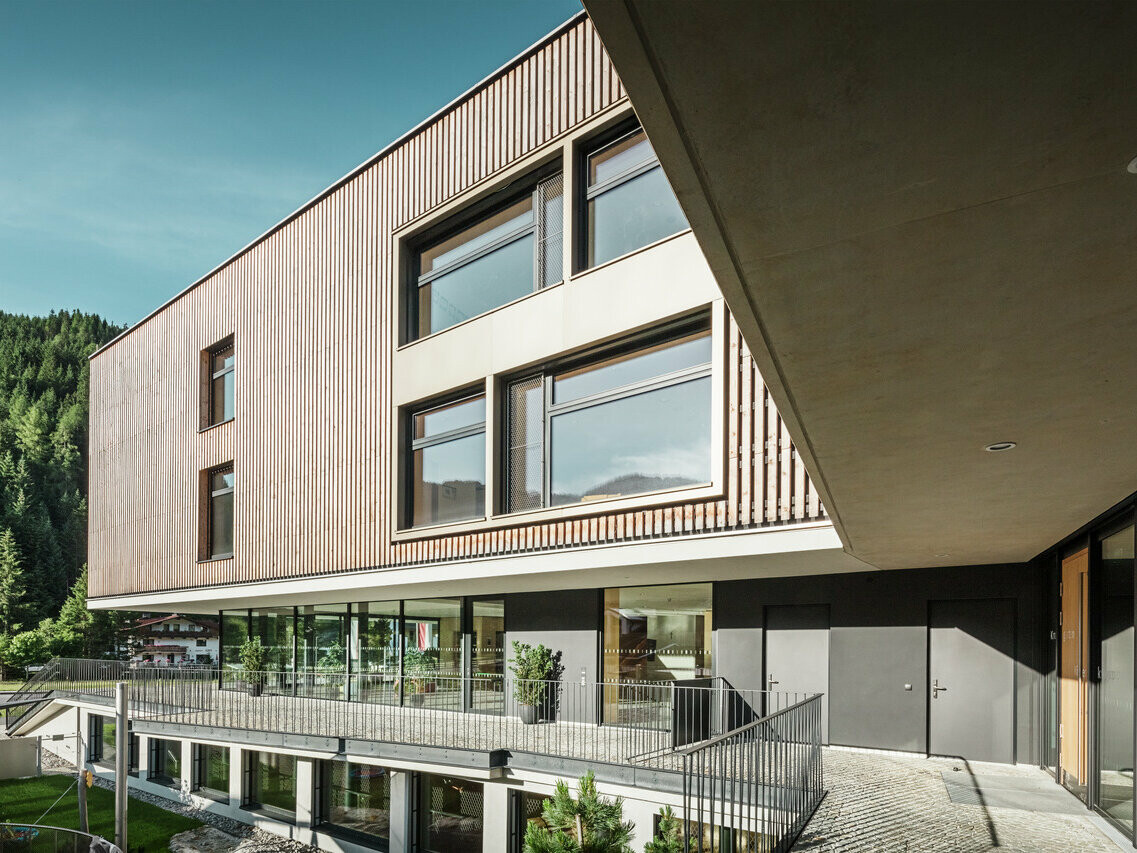 Ein mehrstöckiges Gebäude in moderner Architektur mit einer Kombination aus Holz- und Aluminiumfassade, in einer alpinen Umgebung mit bewaldeten Hügeln im Hintergrund. Das Gebäude hat große Fenster, die viel Licht hereinlassen, und eine überdachte Terrasse, die den Eingang hervorhebt.