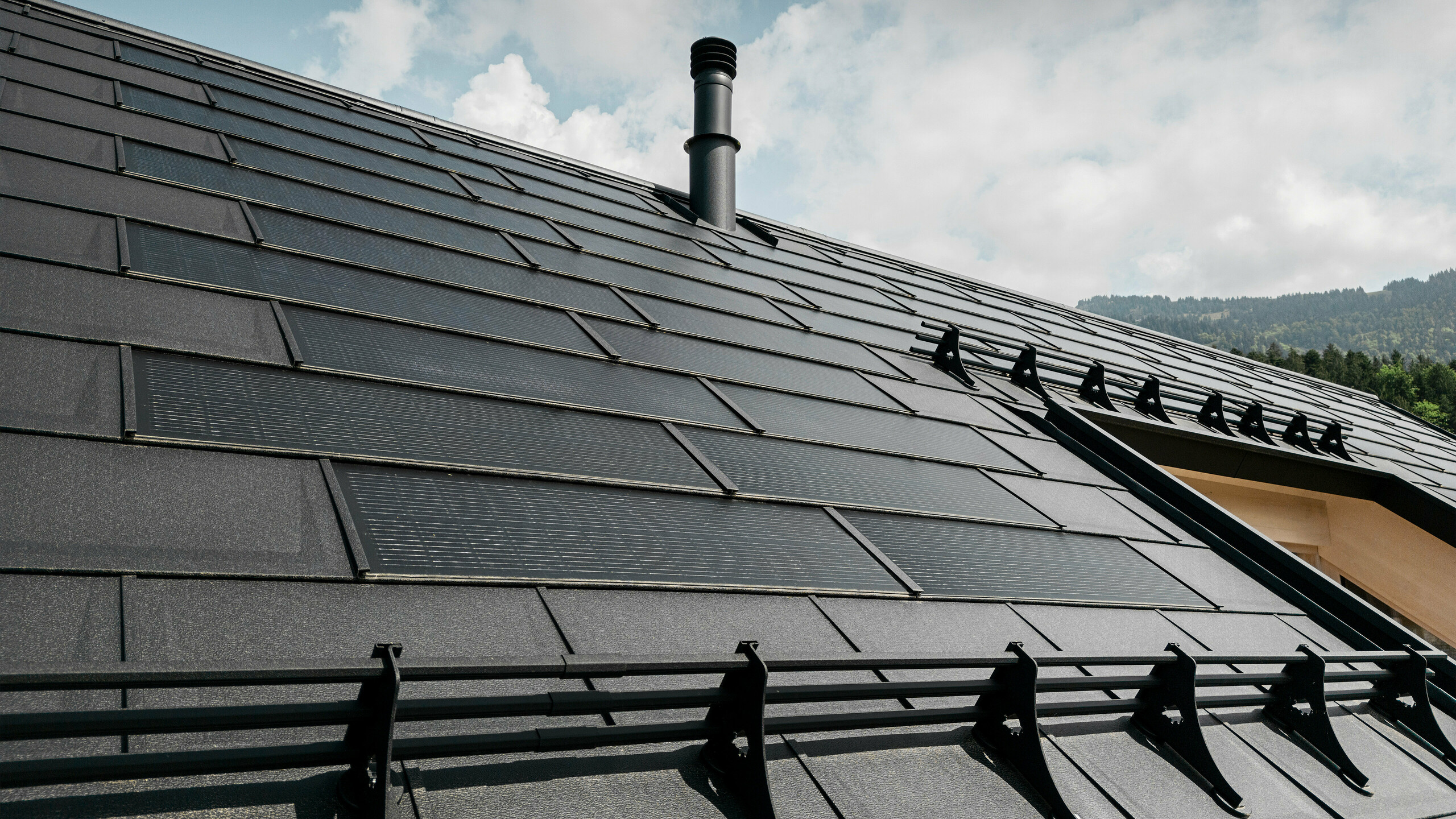 Bližnji posnetek učinkovite solarne strešne plošče PREFA, ki je brezhibno vgrajena v črno streho iz robustnih strešnih plošč PREFA R.16. Kombinacija trajnostne proizvodnje energije in trpežne aluminijaste strehe ponuja ekološko in estetsko strešno rešitev, ki se harmonično zliva s pokrajino.