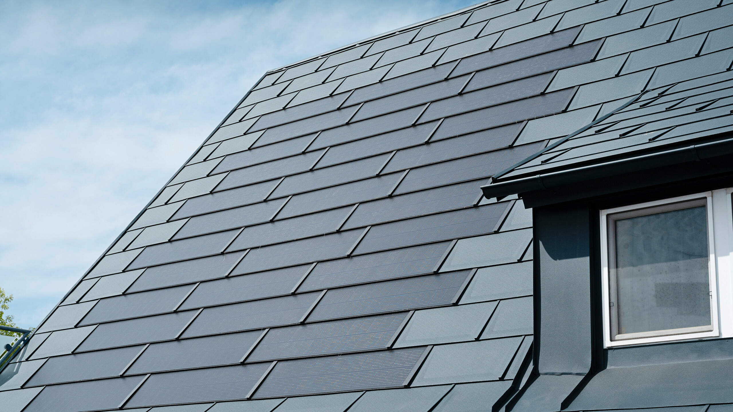 Podroben pogled na prenovljeno streho enodružinske hiše v Leondingu s solarnimi strešnimi ploščami PREFA v antracitni barvi. Od blizu so vidne visokokakovostne aluminijaste plošče PREFA, ki omogočajo brezhibno vgradnjo solarnih modulov. Ta trajna in na vremenske vplive odporna rešitev ponuja trajnosten in energetsko učinkovit način pridobivanja električne energije. Slika ponazarja estetsko in funkcionalno kakovost uporabljenih materialov. Na desni strani slike je pod mansardnim oknom vidno belo okno.