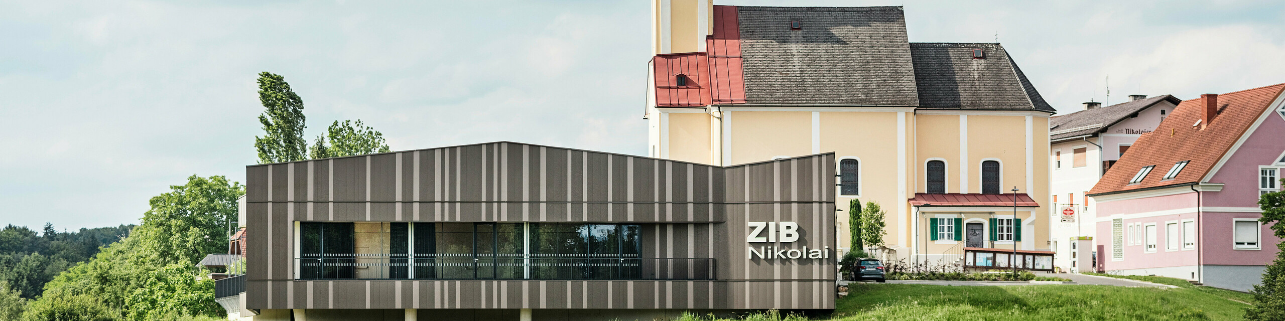 Prireditveni center ZIB Nikolai v St. Veitu na Južnem Štajerskem z moderno, dinamično aluminijasto fasado v rjavo-bež-sivih tonih, v ospredju zelena trata, v ozadju cerkev sv. Nikolaja z rumeno fasado in rdečo streho.