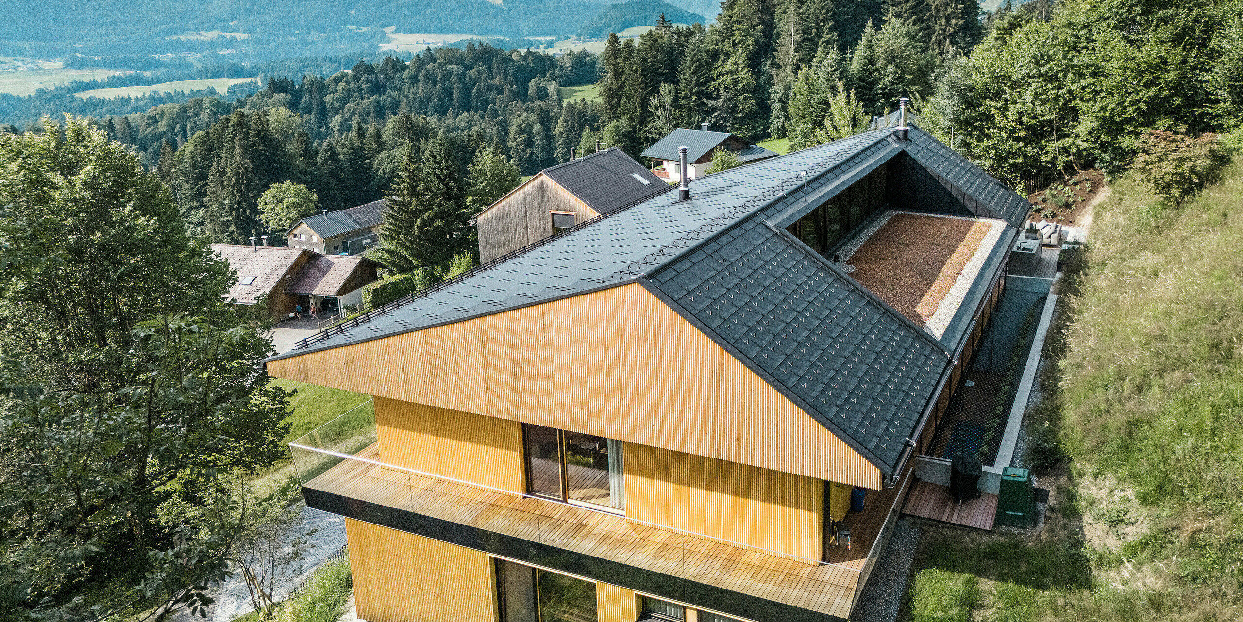 Pogled na leseno fasado enodružinske hiše v kraju Egg v Vorarlbergu. Stavba je bila obložena s solarnimi strešnimi ploščami PREFA in strešnimi ploščami R.16 v črni barvi P.10. Sodobna aluminijasta strešna konstrukcija ima harmonično vključeno strešno ložo na zadnjem delu stavbe. Naravna lesena fasada se s slikovito gorsko panoramo v ozadju odlično zliva z alpsko pokrajino, robustna aluminijasta streha pa zagotavlja trajno zaščito.