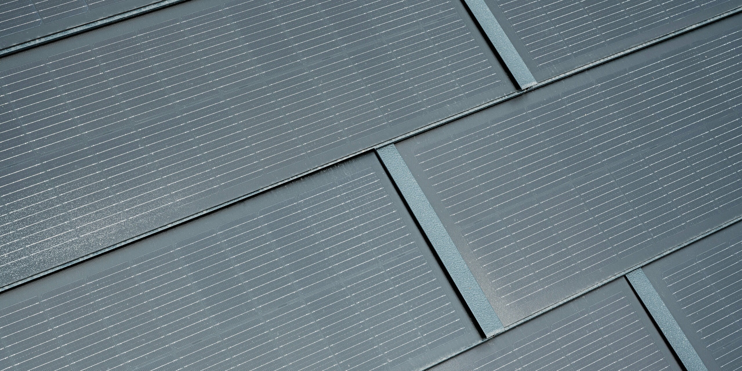 Podroben pogled PREFA solarnih strešnih plošč v antracitni barvi. Fine linije sončnih celic so jasno vidne in se harmonično prilegajo celotnemu dizajnu. Ta privlačna integracija solarne tehnologije v strešno kritino združuje funkcionalnost s sodobno estetiko in zagotavlja okolju prijazen vir energije za stavbe.