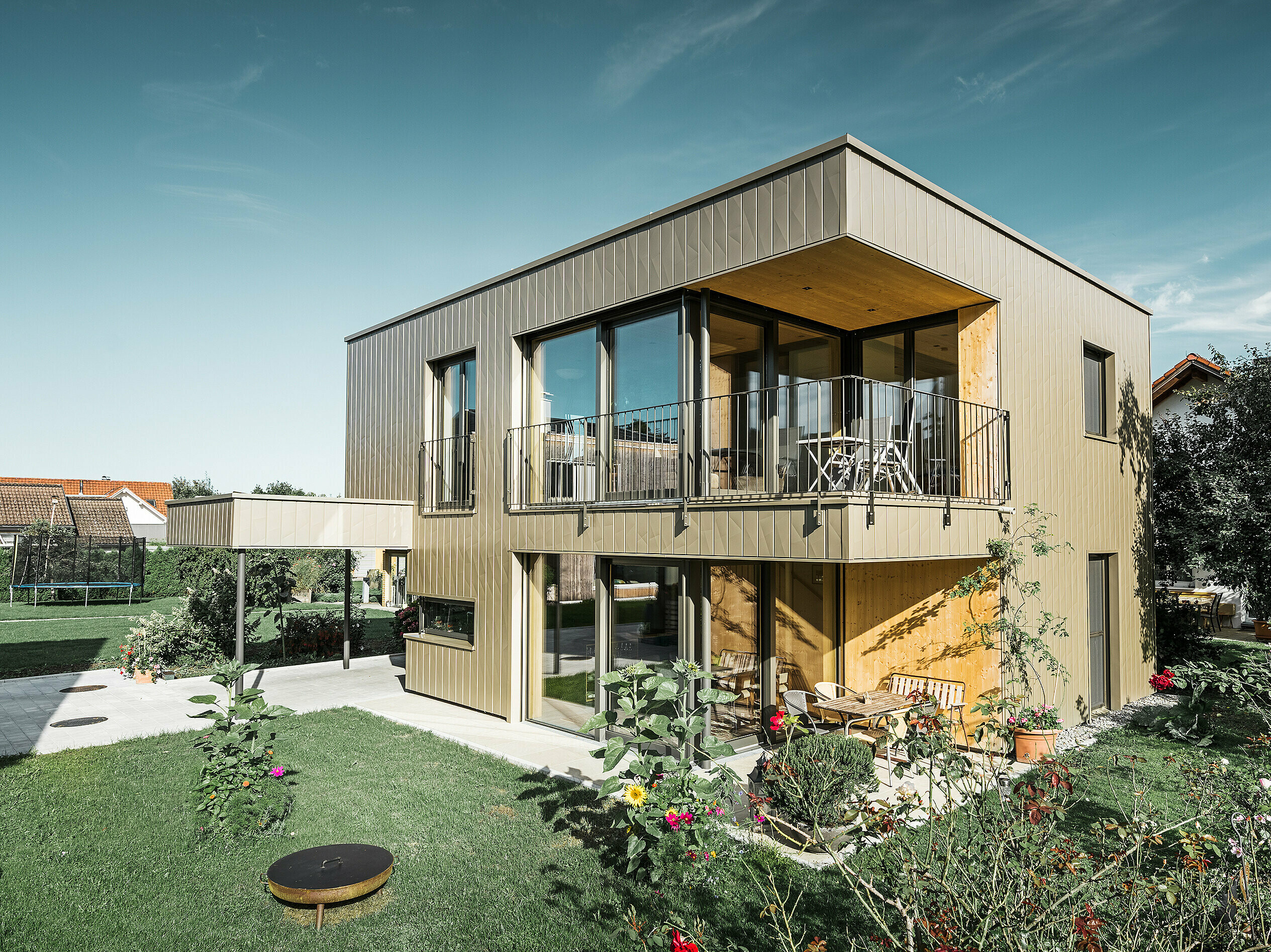 Moderner Neubau mit Flachdach und großen Fensterflächen; Der Großteil der Fassade ist mit vertikalen Alu-Fassaden-Paneelen von PREFA, Siding.X, in Bronze verkleidet. Man erhält auch einen Blick auf den rundum liegenden Garten.