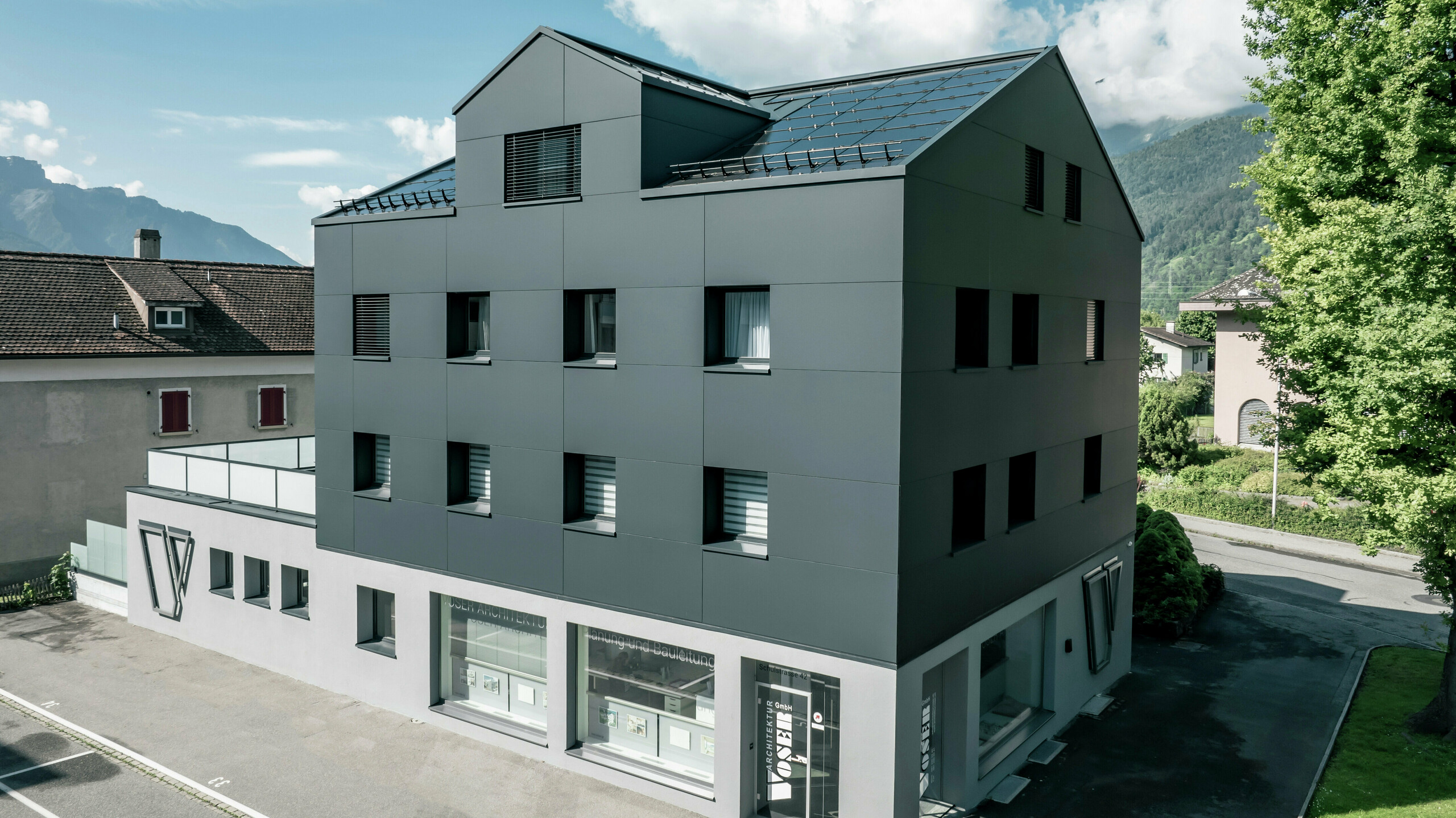 Modernisiertes Wohn- und Geschäftsgebäude nach der Fassadensanierung mit PREFABOND Aluminium Verbundplatten in Anthrazit. Die klare, elegante Linienführung der neuen Fassade verleiht dem Gebäude ein modernes Erscheinungsbild. Im Erdgeschoss befindet sich das Architekturbüro Voser. Die Umgebung mit Bäumen und Bergen rundet die harmonische Optik ab.