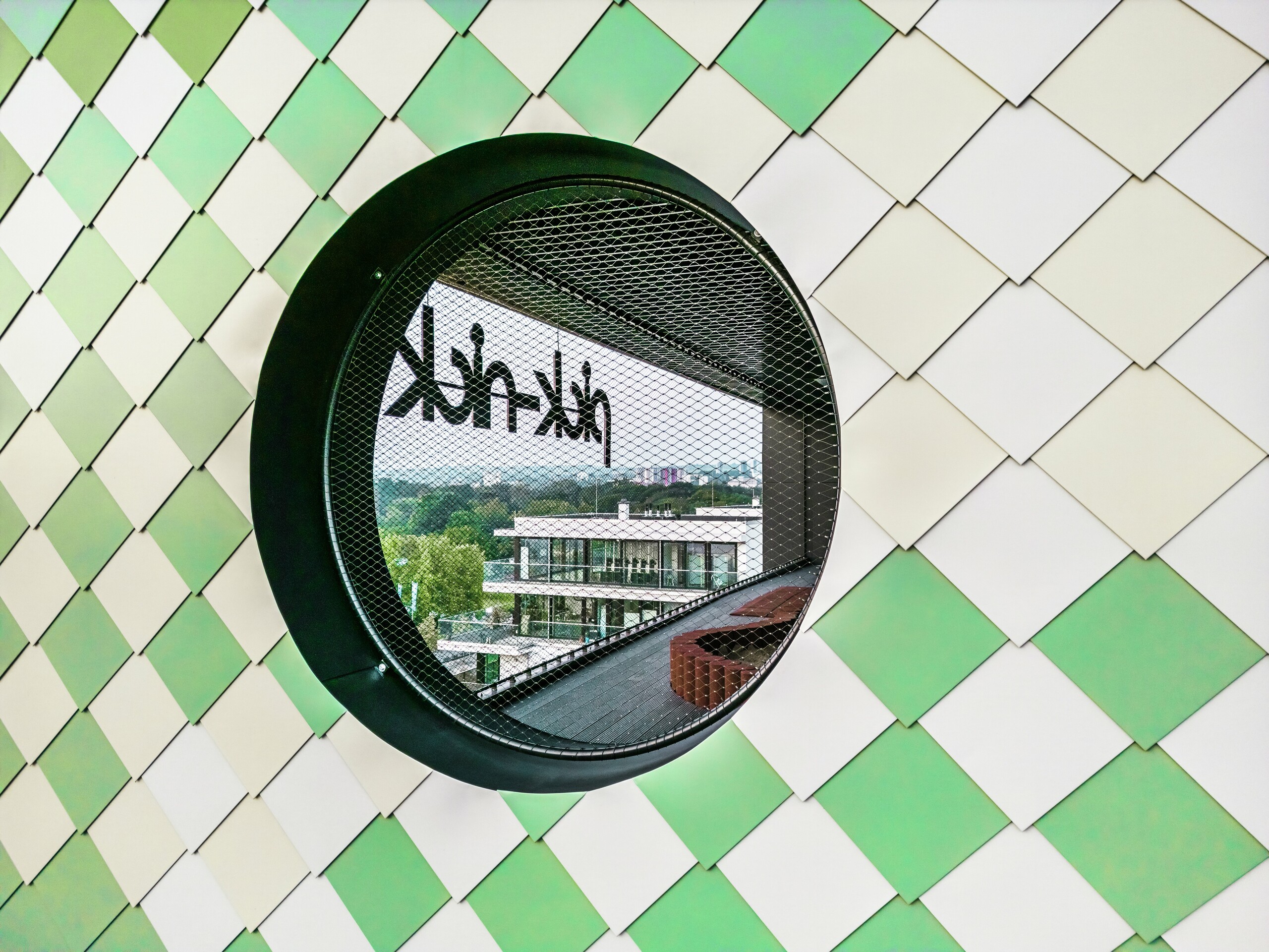 Detailansicht der PREFA Aluminium-Fassade am "Pick-Nick" Wohn- und Geschäftsgebäude in Dresden. Im Mittelpunkt steht ein rundes Fenster mit schwarzem Rahmen und integriertem Gitter. Bei einem Blick durch das runde Fenster ist der spiegelverkehrte Schriftzug "Pick-Nick" zu erkennen. Die Fassade vereint Design, Funktionalität und nachhaltigen Schutz in perfekter Harmonie.