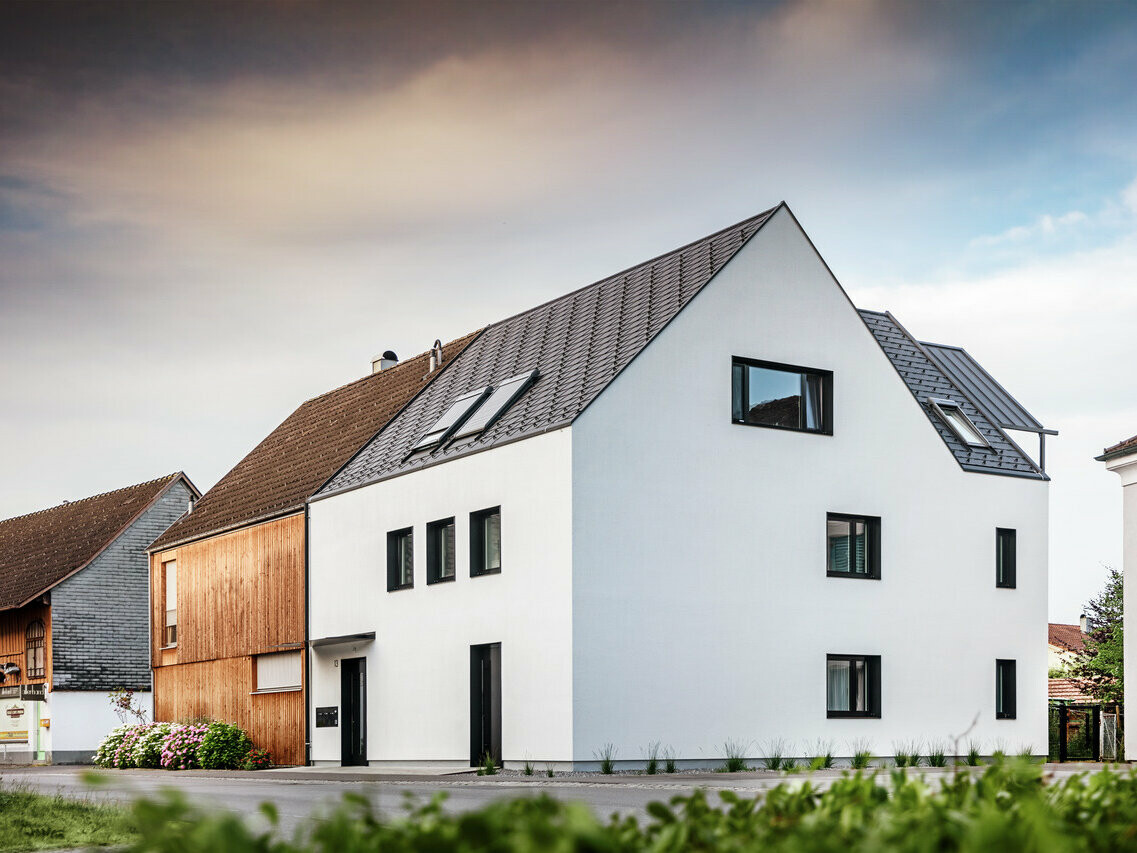 Ansicht eines modernen Mehrfamilienhauses mit einem sattelförmigen PREFA Dach aus Dachschindeln DS19 und Prefalz in Schwarz. Das Haus hat eine strahlend weiße Fassade mit asymmetrisch angeordneten schwarzen Fensterrahmen, die einen markanten Kontrast bieten. Im Vordergrund ist eine gepflegte Grünfläche zu sehen, die das moderne Design des Hauses ergänzt.